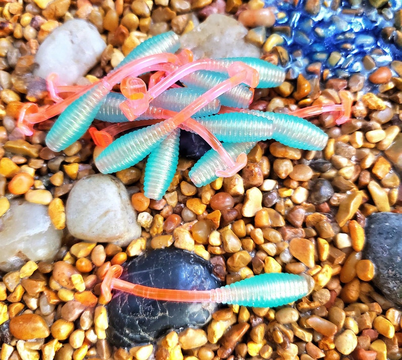 May include: A close-up of a pile of pink and blue fishing lures. The lures are made of soft plastic and have a ribbed texture. They are lying on a bed of small, brown rocks.