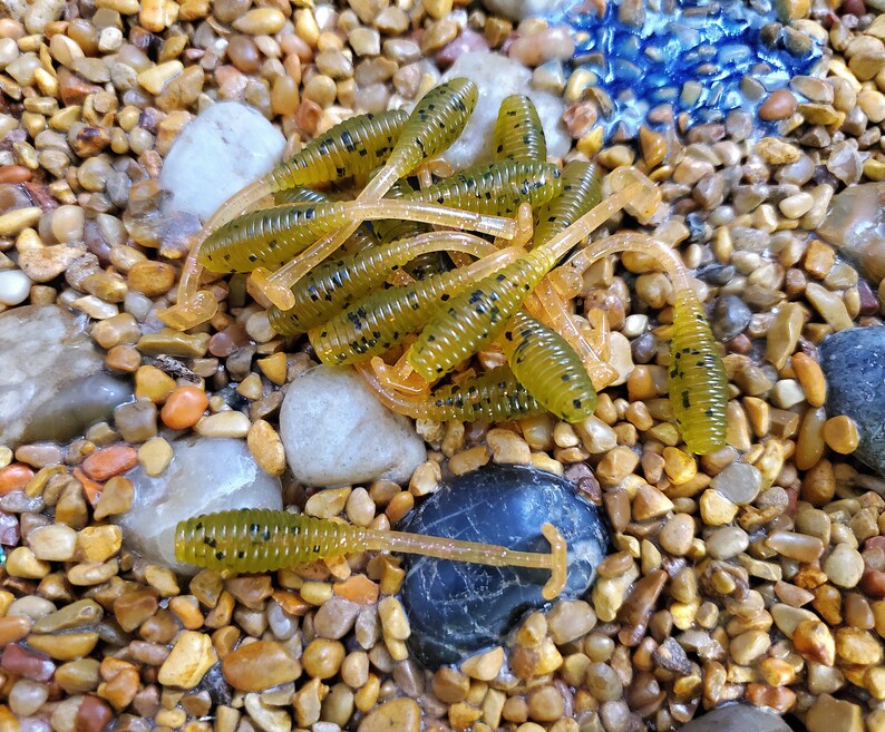 May include: A close-up of a pile of green and brown fishing lures with black speckles. The lures are on a bed of small, smooth, brown and tan stones.