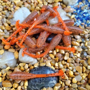 May include: A pile of orange and brown soft plastic fishing lures with black flecks. The lures are shaped like worms and have a curved tail.