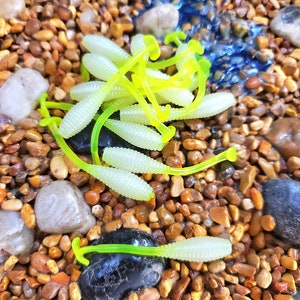May include: A close-up of a pile of bright green and yellow plastic fishing lures. The lures are shaped like worms and are scattered on a bed of small brown rocks.