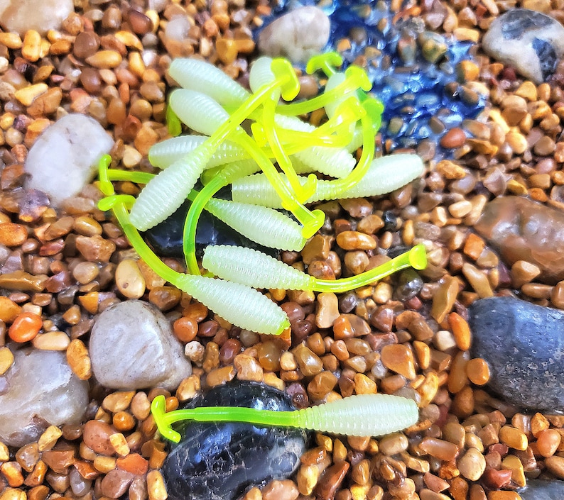 May include: A close-up of a pile of bright green and yellow plastic fishing lures. The lures are shaped like worms and are scattered on a bed of small brown rocks.