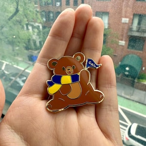 May include: A brown bear enamel pin with a gold outline. The bear wears a blue and yellow scarf and holds a blue flag with "CAL" printed on it. The pin is held in a hand.