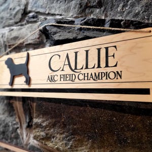 May include: A rectangular light-colored wooden sign featuring a black silhouette of a dog on the left side. The text CALLIE is printed in large black letters above the text AKC FIELD CHAMPION. The sign includes a horizontal slot and twine.