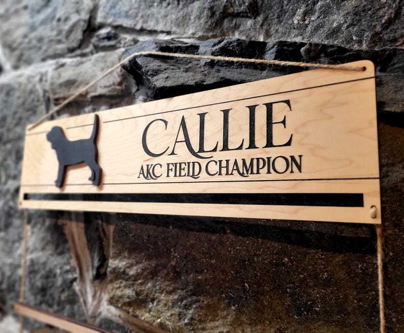 May include: A rectangular light-colored wooden sign featuring a black silhouette of a dog on the left side. The text CALLIE is printed in large black letters above the text AKC FIELD CHAMPION. The sign includes a horizontal slot and twine.