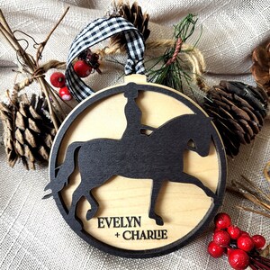 May include: A wooden Christmas ornament featuring a black silhouette of a rider on a horse. The ornament is personalized with the names "Evelyn + Charlie".