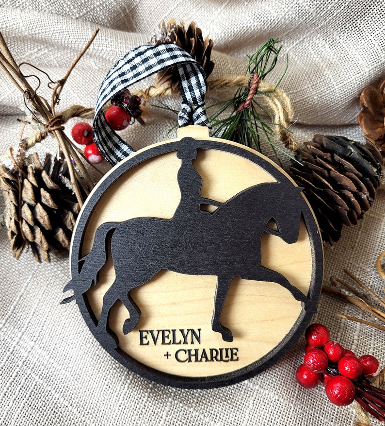 May include: A wooden Christmas ornament featuring a black silhouette of a rider on a horse. The ornament is personalized with the names "Evelyn + Charlie".
