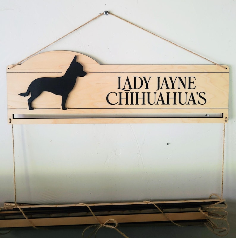 May include: A light-colored wooden wall sign featuring a black silhouette of a chihuahua and the black text LADY JAYNE CHIHUAHUA'S. The sign includes a horizontal slot and is suspended by thin twine, with a matching wooden bar hanging below.