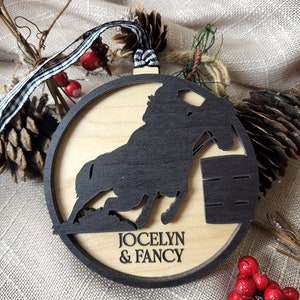 May include: A wooden Christmas ornament featuring a black silhouette of a horse jumping over a barrel. The ornament is personalized with the names "Jocelyn & Fancy".