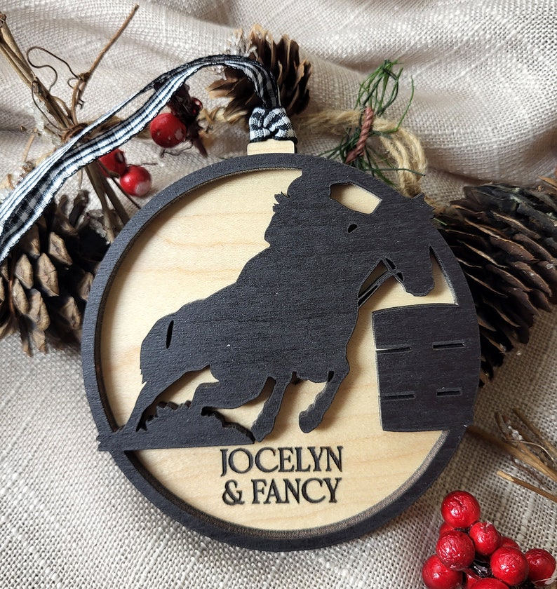 May include: A wooden Christmas ornament featuring a black silhouette of a horse jumping over a barrel. The ornament is personalized with the names "Jocelyn & Fancy".