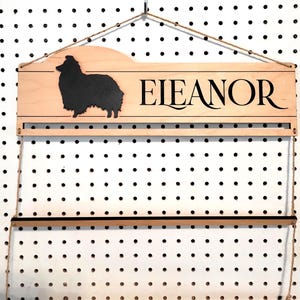 May include: A light-colored wooden wall-mounted display rack featuring a black silhouette of a dog on the left and the name "EIEANOR" in black lettering on the right. The rack includes a horizontal bar and is suspended by thin twine.