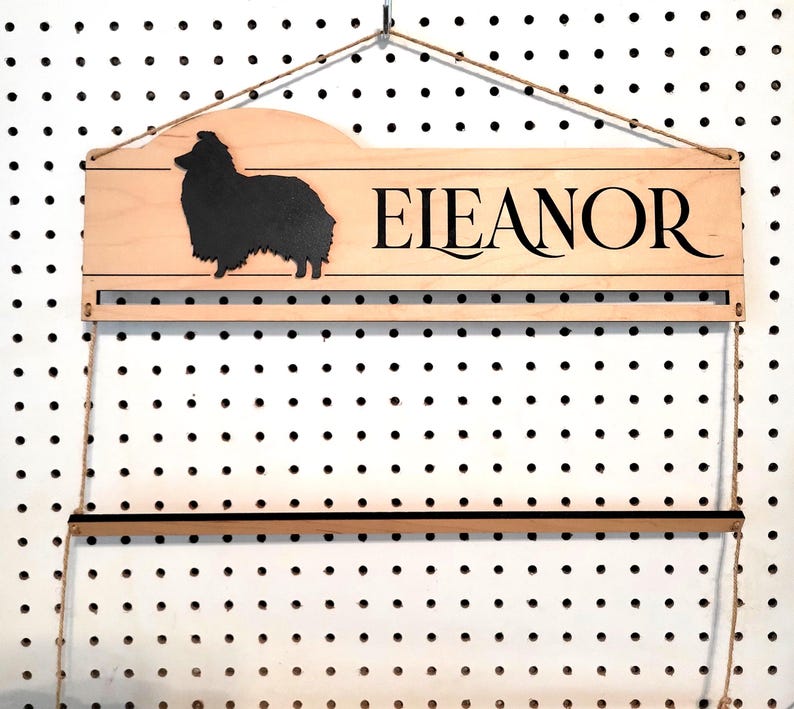 May include: A light-colored wooden wall-mounted display rack featuring a black silhouette of a dog on the left and the name "EIEANOR" in black lettering on the right. The rack includes a horizontal bar and is suspended by thin twine.