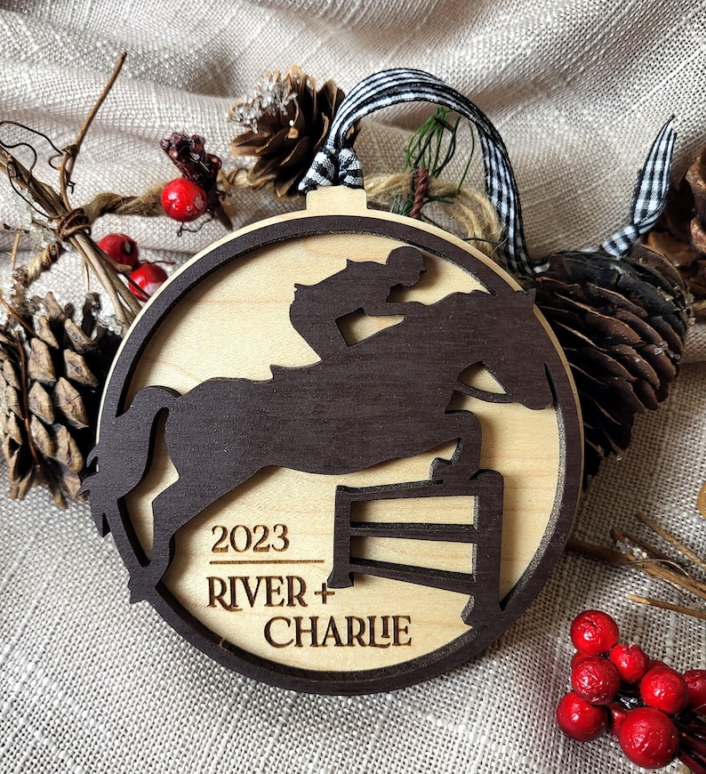 May include: A wooden Christmas ornament featuring a silhouette of a horse jumping over a hurdle. The ornament is decorated with the year 2023, the names "River + Charlie", and a brown woodgrain finish.