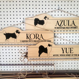 May include: Three light-colored wooden wall signs with hanging twine. Each sign features a black silhouette of a dog and engraved text: AZULA NORTHSTARS BLUE INFERNO, KORA CAIDERSONGS BREATH OF THE WILD, and YUE MYSTIC WINDS SPIRIT OF THE MOON.
