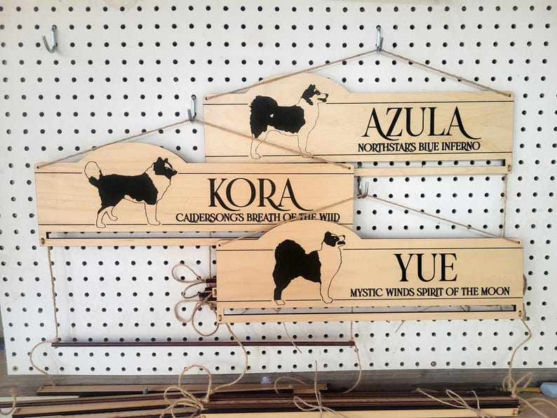 May include: Three light-colored wooden wall signs with hanging twine. Each sign features a black silhouette of a dog and engraved text: AZULA NORTHSTARS BLUE INFERNO, KORA CAIDERSONGS BREATH OF THE WILD, and YUE MYSTIC WINDS SPIRIT OF THE MOON.