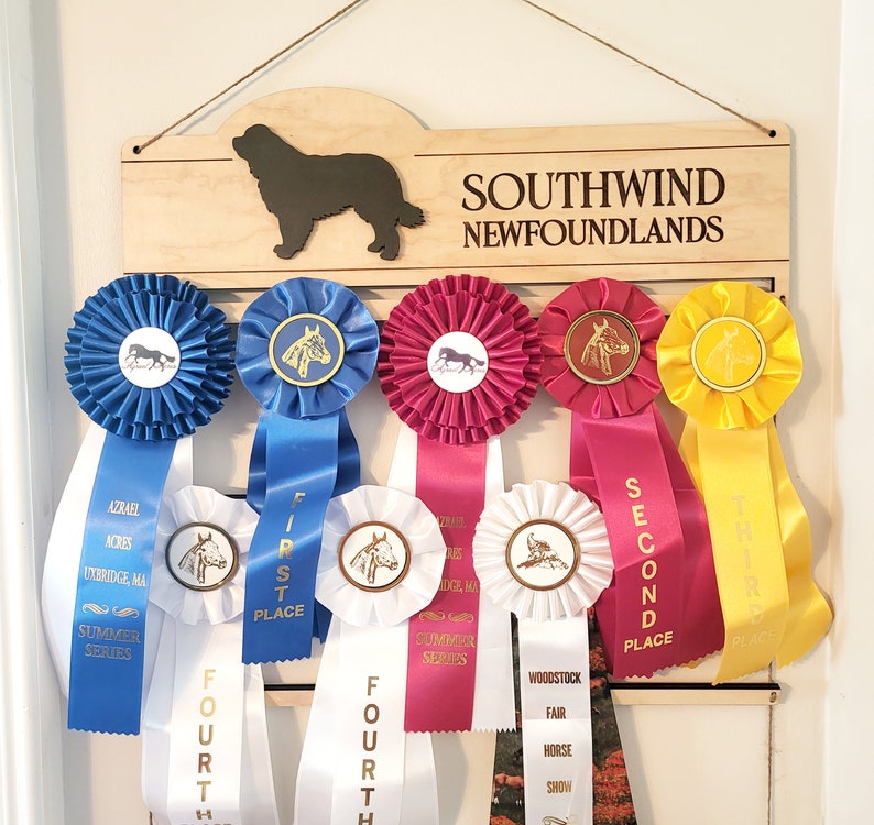 May include: A light-colored wooden display board with a cutout of a Newfoundland dog and the text SOUTHWIND NEWFOUNDLANDS. It holds several colorful award ribbons in blue, magenta, and yellow, featuring various horse and dog show graphics.