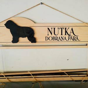 May include: A light-colored wooden wall display featuring a black silhouette of a dog on the left and the black text NUTKA DOBRANA PARA on the right. The piece includes a horizontal bar and a lower section suspended by twine for hanging items.