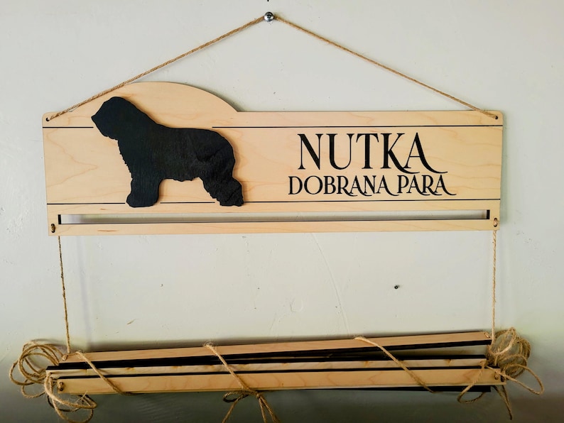 May include: A light-colored wooden wall display featuring a black silhouette of a dog on the left and the black text NUTKA DOBRANA PARA on the right. The piece includes a horizontal bar and a lower section suspended by twine for hanging items.