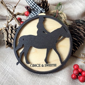 May include: A round wooden ornament with a black silhouette of a person riding a horse. The ornament is decorated with the text "GRACE & SWEETIE".