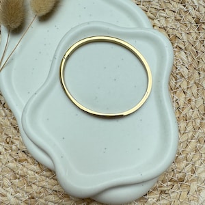 Könnte beinhalten: Ein goldfarbenes Armreif-Armband liegt auf einer gesprenkelten, cremefarbenen Keramikschale. Die Schale steht auf einer gewebten, beigefarbenen Oberfläche. Getrocknete, flauschige beige Pflanzen sind auf der Schale platziert.