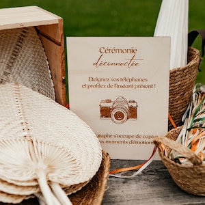 Peut inclure: Une pancarte en bois avec le texte "Cérémonie déconnectée", qui signifie "Cérémonie débranchée". La pancarte comprend également le texte "Eteignez vos téléphones et profitez de l'instant présent !", qui signifie "Éteignez vos téléphones et profitez du moment présent !". La pancarte est sur une table en bois avec un panier de rubans colorés et une boîte d'éventails en osier.