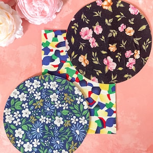Può includere: Una collezione di sottobicchieri in tessuto e un pezzo rettangolare con motivi geometrici colorati. Un sottobicchiere è blu navy con fiori bianchi, un altro è nero con fiori rosa e il terzo ha un design astratto colorato. Rose rosa sullo sfondo.