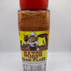 May include: A clear plastic jar with a red lid filled with a red and orange spice blend. The label is yellow with a cartoon chef and the text "SAZON RANCHO DON FLOR" and "Sazon Para Langostinos, Camaron y Cucarachas".