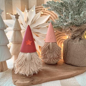 May include: Two decorative gnome figurines with red and pink conical hats, each adorned with a star. The gnomes have beige bodies and long, fringed beards. They sit on a wooden board, with a small Christmas tree and other festive decorations in the background.