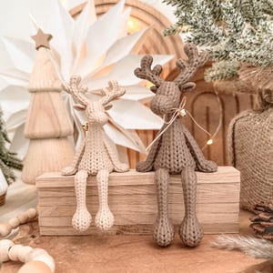 May include: Two crocheted reindeer figurines sitting on a wooden block. The left reindeer is cream-coloured, and the right reindeer is brown. Each reindeer features a textured knit pattern, long dangling legs, and a small gold bell tied around its neck.