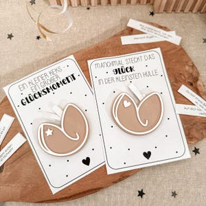 May include: Two fortune cookie-shaped decorations with white frosting details and a star or heart design, presented on cards with German text. The cards are on a wooden surface, with additional fortune slips.