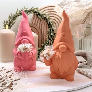 Könnte beinhalten: Zwei dekorative Zwergfiguren. Eine ist leuchtend rosa, die andere warm orange. Jeder Zwerg hält einen kleinen Topf mit getrockneten weißen Blumen. Der rosa Zwerg hat ein Herzdetail, während der orangefarbene eine Blume auf seinem Hut hat. Sie stehen auf einem gestreiften Tablett.