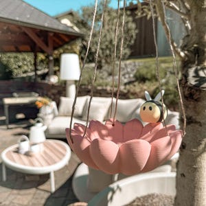 Op de afbeelding: Een hangende roze bloemvormige vogelvoeder met een klein bijenfiguur op de rand. De voederbak is opgehangen met touw en staat tegen een vage buitenachtergrond met een terras en bomen.