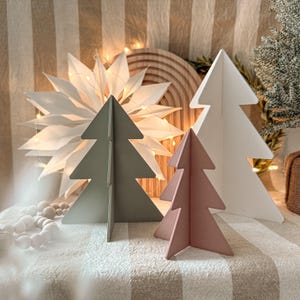 Puede incluir: Una exhibición festiva con tres árboles de Navidad de madera en tonos verdes y rosas, junto a una gran estrella de papel blanco y luces decorativas. Los árboles están sobre una tela a rayas.