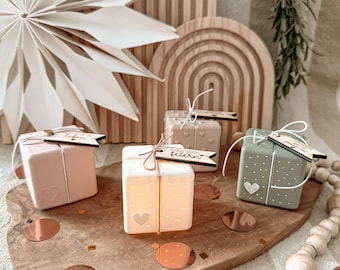 May include: Four square, pastel-colored gift boxes with heart and dot designs, each with a small wooden tag tied with twine. The boxes are arranged on a wooden board with copper confetti. A large white paper star and wooden arch decor are in the background.