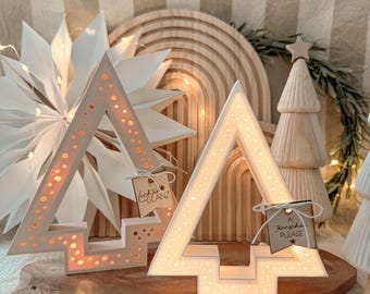 May include: Two white, illuminated Christmas tree-shaped decorations with cut-out dot patterns. Each tree has a tag attached. Other wooden Christmas decorations are in the background, including a star, a tree, and a wooden rainbow.