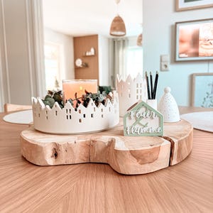 Puede incluir: Un centro de mesa decorativo sobre una losa de madera. Presenta un portavelas de cerámica blanca con diseño de pueblo, una vela encendida y vegetación. También hay un pequeño letrero verde en forma de casa con texto.