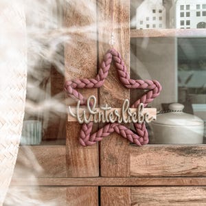 Könnte beinhalten: Ein sternförmiger Wandbehang aus geflochtenem, altrosa Stoff. Über die Mitte ist eine horizontale Holzleiste angebracht, auf der das Wort Winterliebe in einer kursiven, silberfarbenen und strukturierten Schriftart zu sehen ist.