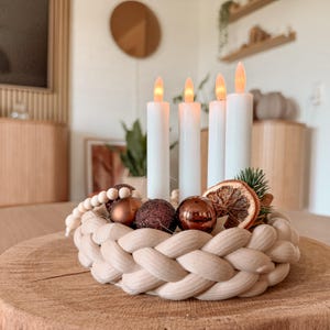 Könnte beinhalten: Ein runder, geflochtener Kranz aus beigem Stoff, verziert mit vier weißen LED-Stabkerzen, metallisch bronzefarbenen Kugeln, einer Holzperlenkette und einer getrockneten Orangenscheibe. Die Kerzen haben leuchtende orangefarbene Flammen.
