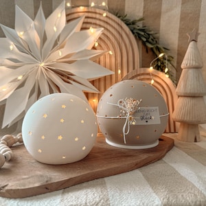Könnte beinhalten: Zwei kugelförmige dekorative Lichter, eines cremefarben und eines beige, mit Sternaussparungen. Das beige Licht hat ein Etikett mit der Aufschrift "WENIG REICHT ZUM Glück". Ein Papierstern, Holzbögen und ein Holzbaum sind im Hintergrund.