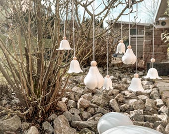 May include: White ceramic bell-shaped ornaments hanging from a tree branch. Each ornament has a wooden bead and is suspended by a thin white string. The ornaments vary in size and shape, creating a whimsical outdoor display.