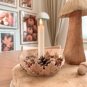 Könnte beinhalten: Eine beige Keramikschale in Form von Ahornblättern, gefüllt mit Tannenzapfen und kleinen Dekorationsobjekten. Eine hohe, cremeweiße LED-Stabkerze steht in der Mitte. Neben der Schale befindet sich eine hohe, strukturierte, hellbraune Pilzfigur.