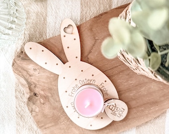 May include: A wooden bunny-shaped candle holder with a pink tea light candle inside. The holder has the text "Frohe Ostern" (Happy Easter) around the edge and a small heart cutout on the bunny's ear.