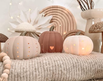Puede incluir: Tres pequeñas luces decorativas con forma de calabaza en tonos beige claro, terracota y blanco. Cada calabaza presenta un tallo verde oscuro y pequeñas aberturas, incluida una forma de corazón en la de color terracota, que emiten una luz cálida.