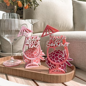 May include: Two pink laser-cut drink decorations with raspberry designs and text. One says "It's pink berry time" and the other says "So schmeckt der Sommer". They sit on a wooden tray with a wine glass.