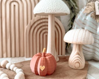 May include: A selection of wooden decorative items. These include a coral-coloured pumpkin with a gold heart, two wooden mushrooms, and a large mushroom with a white knitted cap. A wooden arch and a beaded garland are also visible.