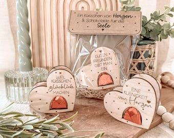 May include: Three wooden heart-shaped boxes with a chocolate inside. Each box has a different message engraved on it: "Weil Kusschen glucklich machen", "Guten Freunden gibt man ein", and "Eine Kleinigkeit von Herzen".