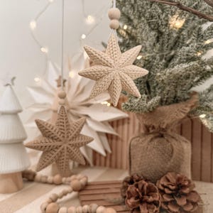 Puede incluir: Dos adornos en forma de estrella de ocho puntas de color beige con un patrón texturizado similar al tejido. Cada estrella cuelga de un cordel blanco con una pequeña cuenta de madera. Las estrellas tienen un acabado mate y forma tridimensional.