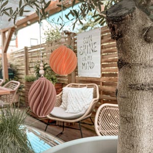 Puede incluir: Dos adornos decorativos esféricos colgantes, en tonos melocotón y malva. Los adornos tienen un diseño en espiral y están suspendidos por una cuerda con cuentas de madera. Un letrero en el fondo dice "SUN SHINE ON MY MIND."