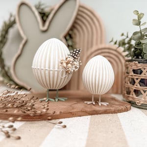 Puede incluir: Dos figuras decorativas de huevos de Pascua. Los huevos son blancos con rayas verticales beige y se apoyan sobre patas verdes y blancas. Un huevo está adornado con un pequeño arreglo floral y una pluma. Los huevos están sobre una tabla de madera.