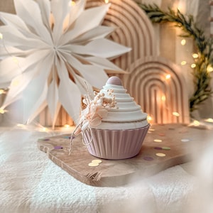 May include: A cupcake-shaped container in pale pink and white, decorated with dried flowers. The container sits on a wooden board with confetti, against a backdrop of a paper star and fairy lights. A decorative item.