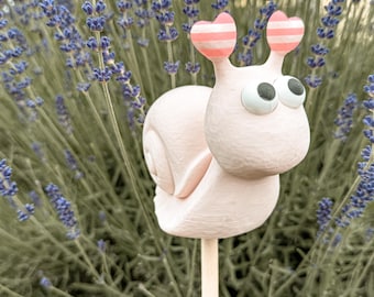 Könnte beinhalten: Ein hellrosa Schnecken-Gartenstecker mit texturiertem Gehäuse und Körper. Die Schnecke hat zwei große, runde, weiße Wackelaugen und zwei Fühler, die mit kleinen, horizontal gestreiften rosa-weißen Herzakzenten auf einem Holzstab versehen sind.